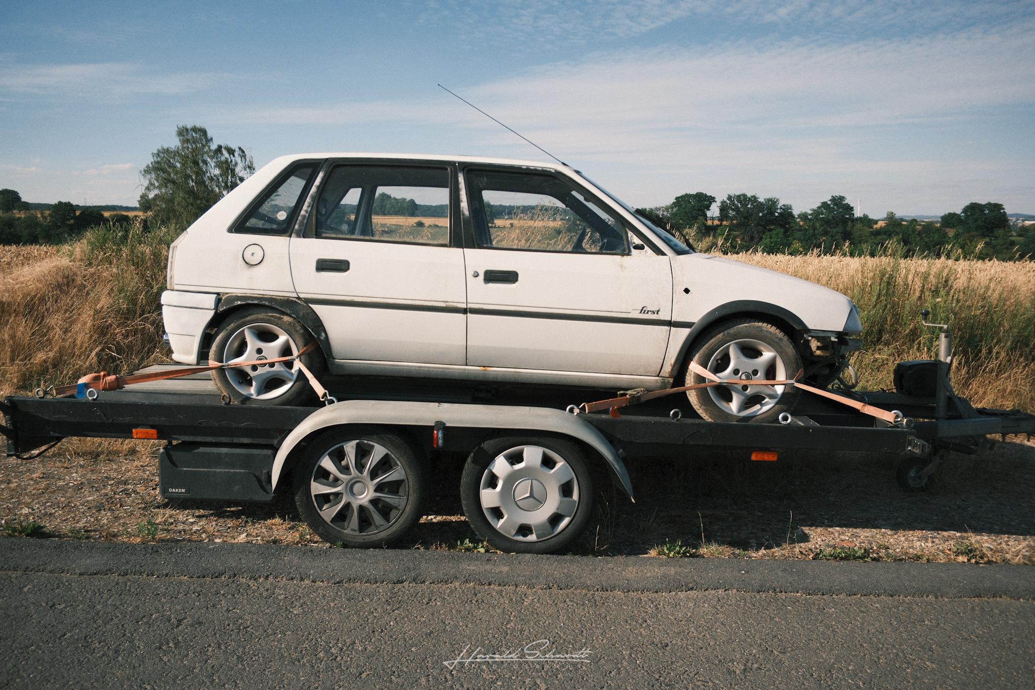 Old white hatchback strapped down on a car trailer parked on the roadside, dry field behind it.