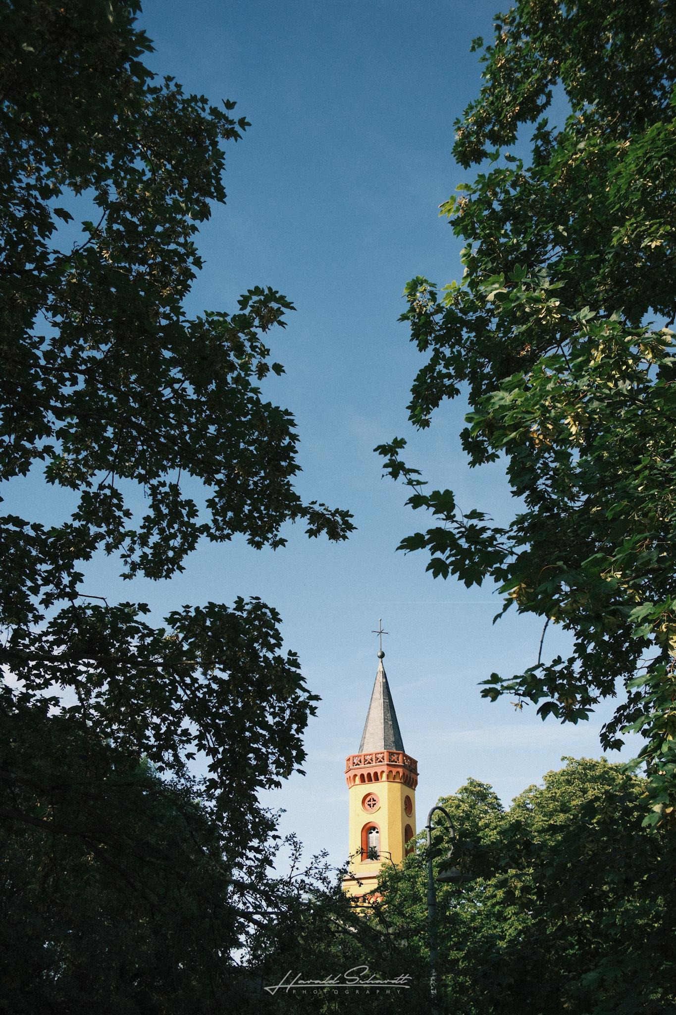 Church tower with a yellow and red facade, spire topped with a cross, framed by tree branches against a blue sky.