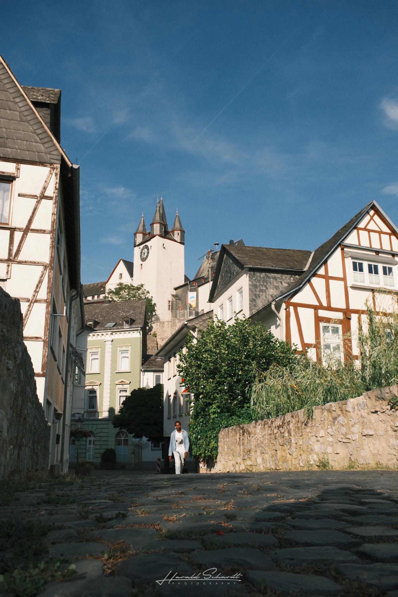 Cobblestone street leading up to Diez Castle's towers, half-timbered houses on both sides, a person walking in the distance.