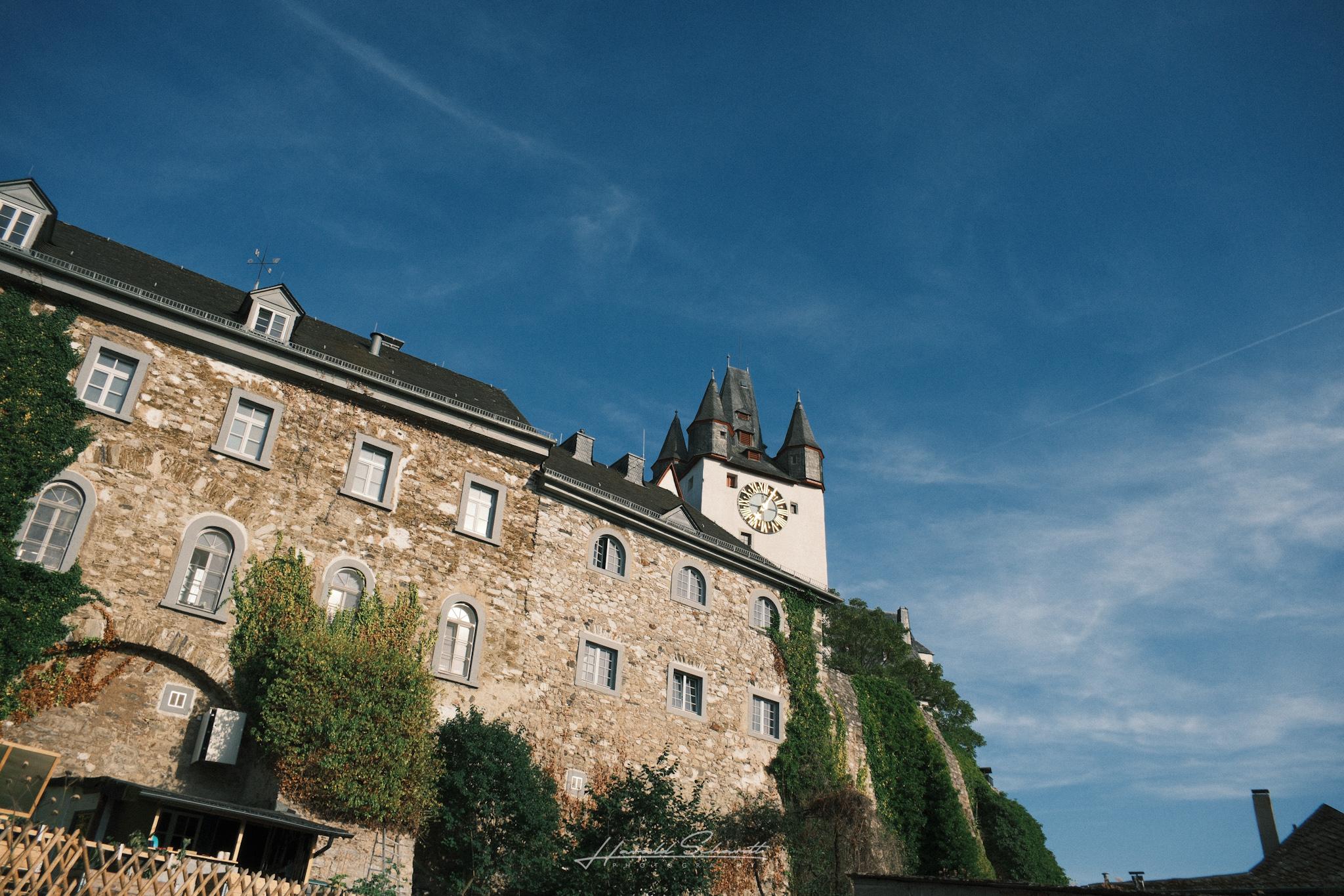 Diez Castle seen from below, stone facade covered in ivy, one of the towers with a clock visible above.
