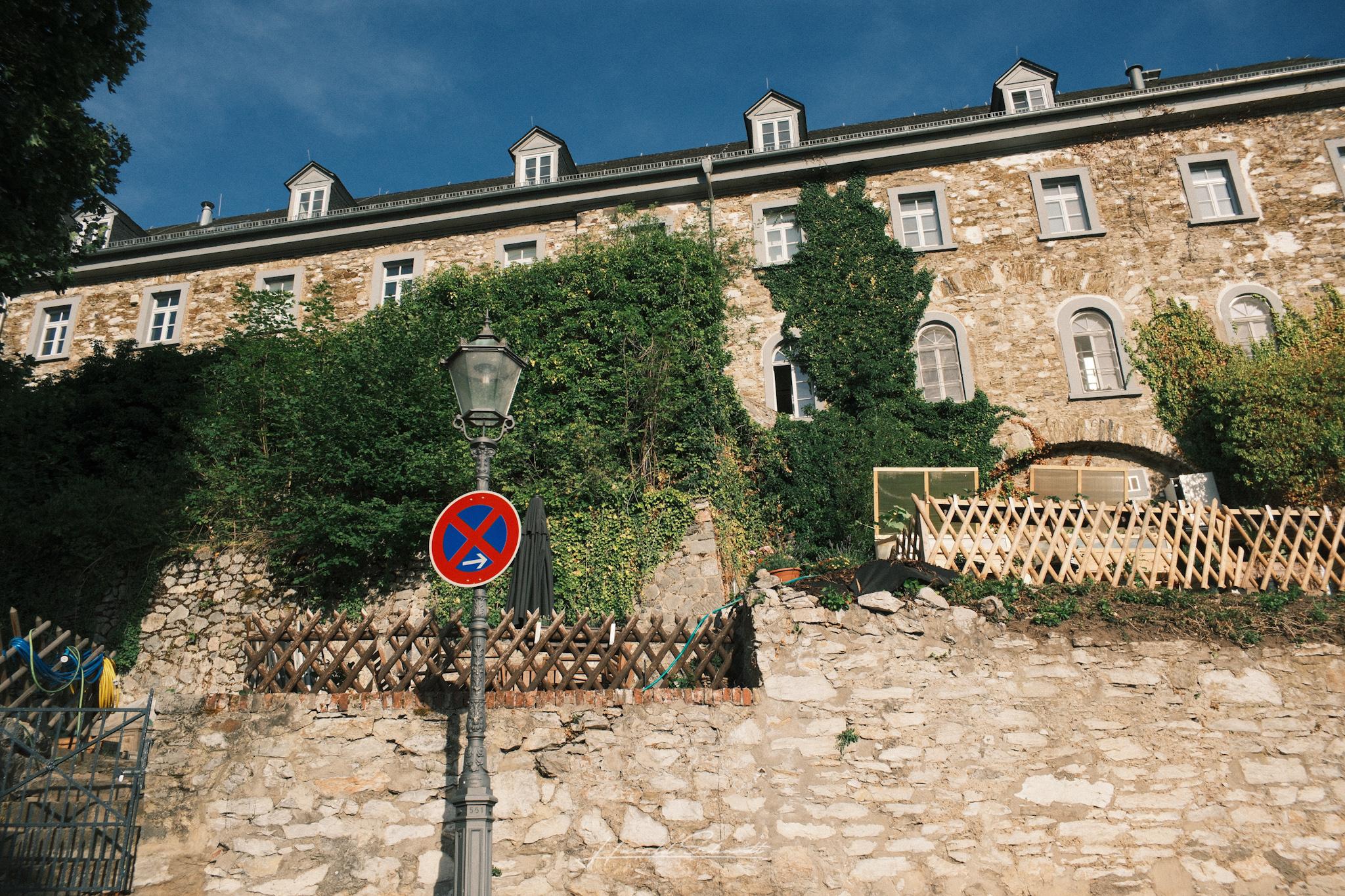 Castle wall covered in ivy, an old lamp post in front, a no parking sign and garden fencing along the path.