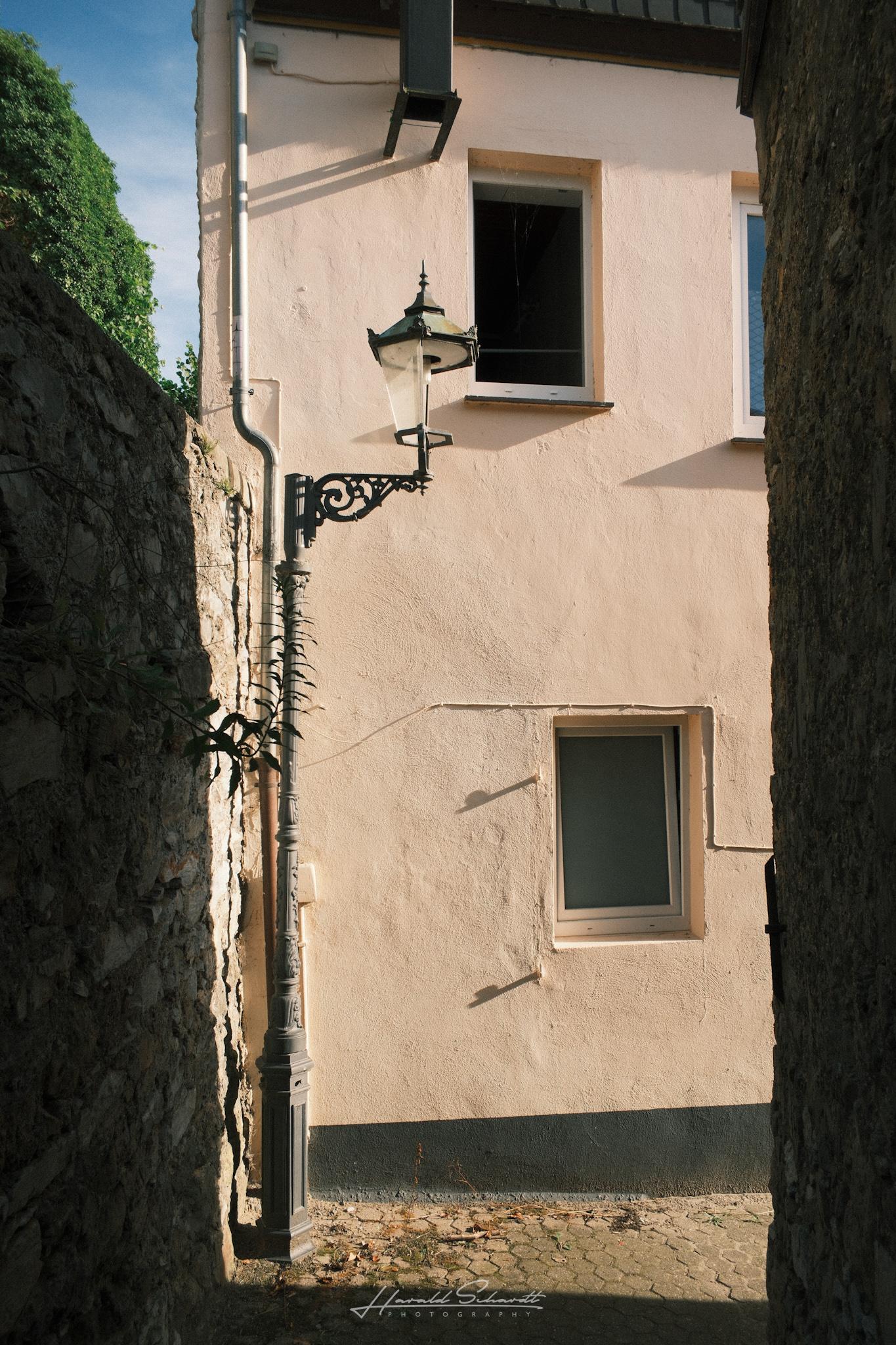 Narrow alley between a stone wall and a plastered house, an old lamp fixture mounted on the facade, strong light and