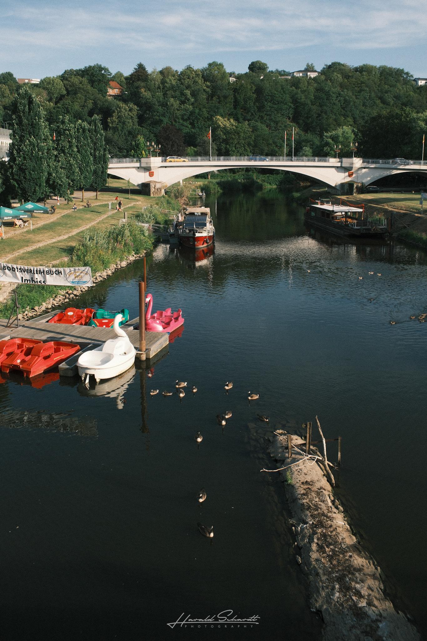 View of the Lahn river from a bridge, swan and flamingo shaped pedal boats docked, a stone bridge and ducks on the water.