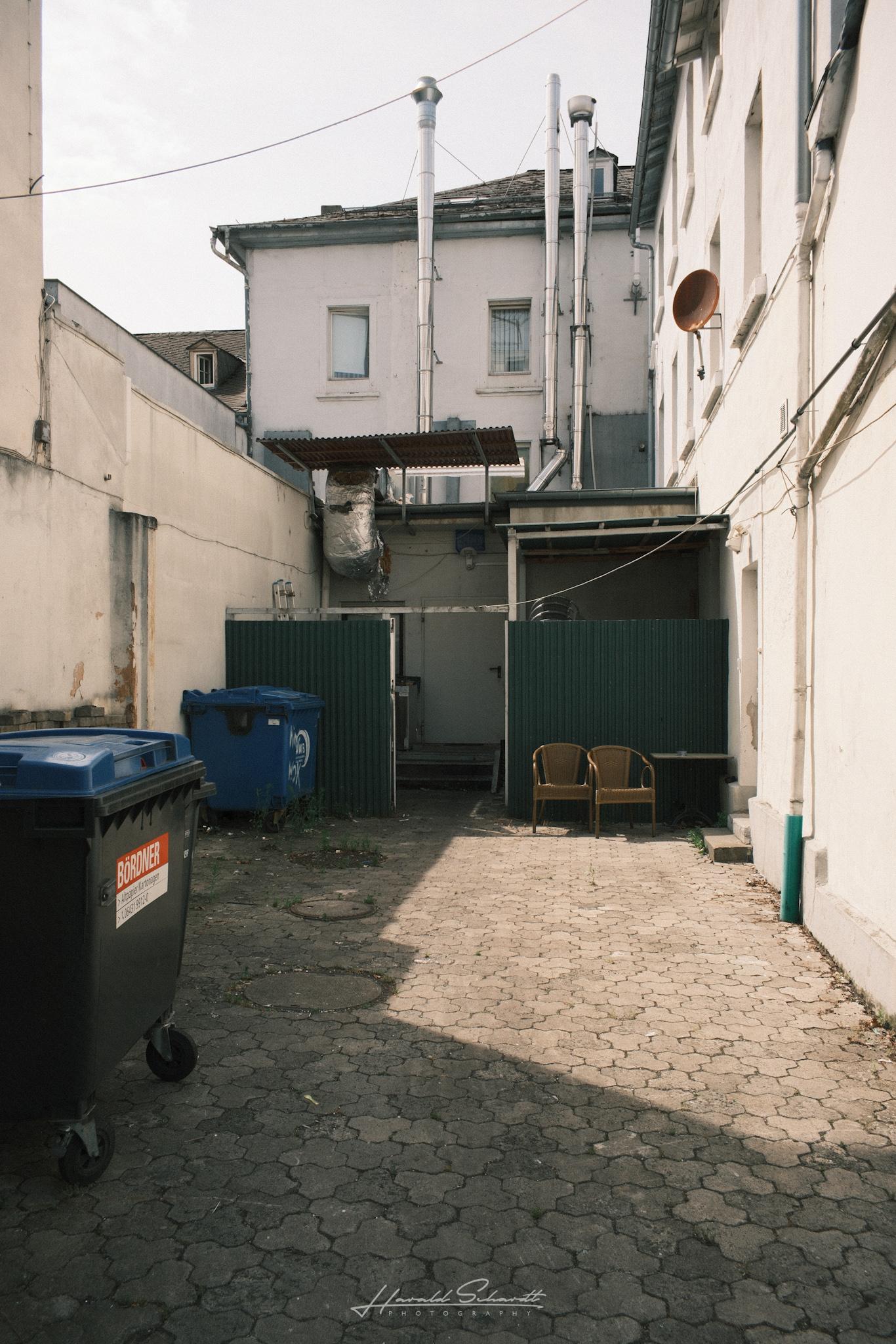 A quiet backyard behind the city center with recycling bins, two old armchairs, and industrial pipes along the buildings.