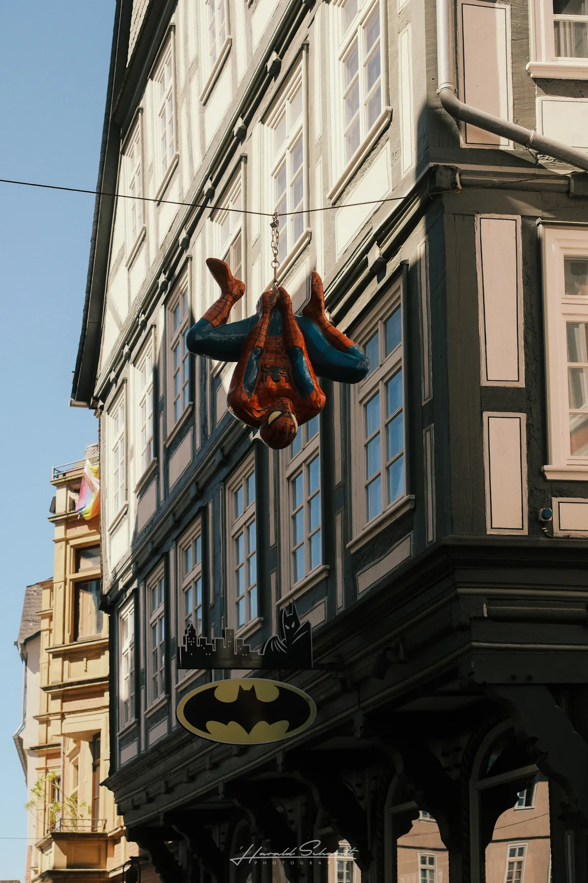A Spider-Man figure hanging upside down from a wire above a Batman shop sign on a half-timbered building in Marburg.