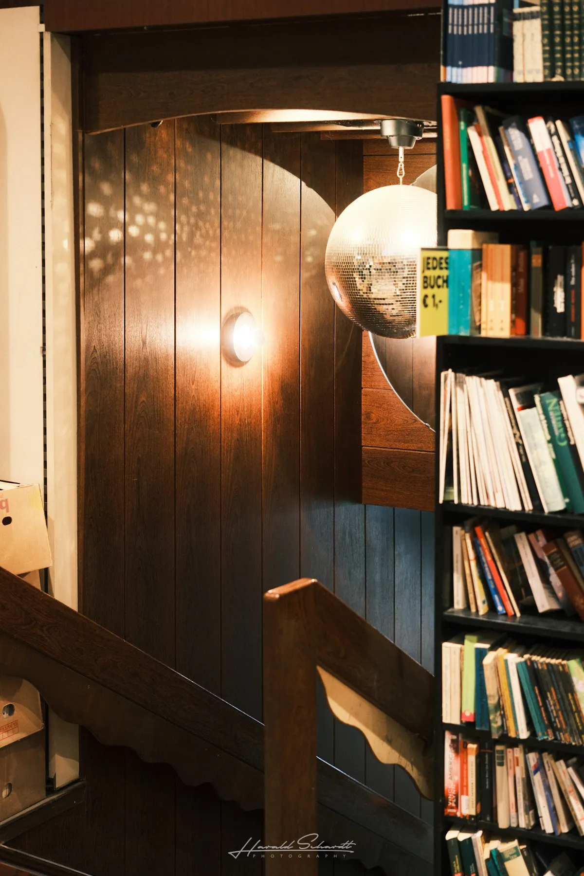 A disco ball hanging at the top of a wooden staircase in a dimly lit second-hand shop, bookshelves to the right.