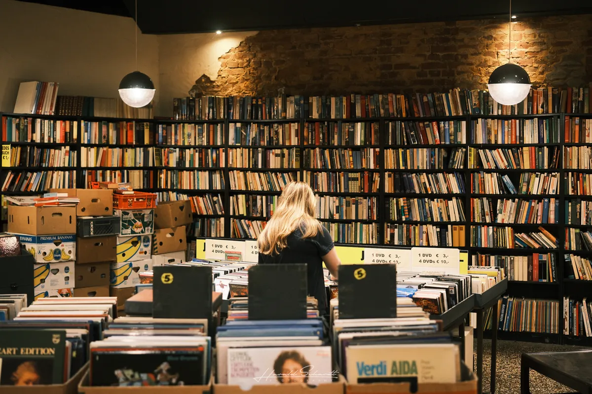Inside a second-hand record and book shop, a woman browsing vinyl crates in front of floor-to-ceiling bookshelves.