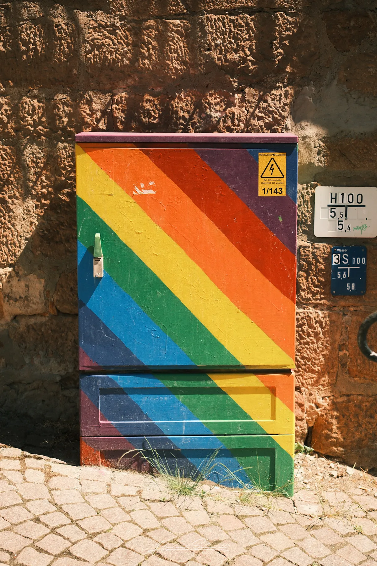 An electrical box painted with a rainbow flag design, set against a rough stone wall on a cobblestone street.