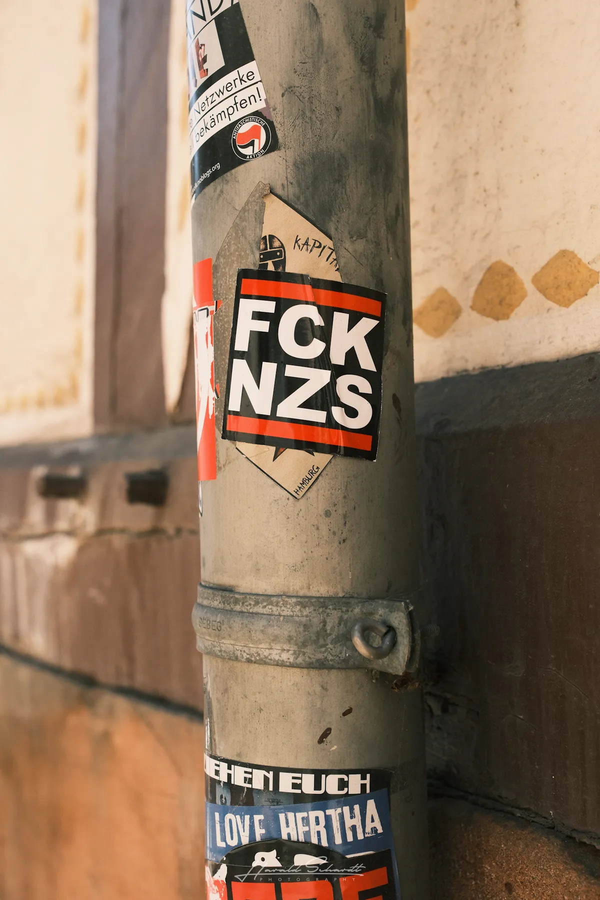 A lamp post covered in political stickers, including a prominent FCK NZS sticker, against a rendered wall.