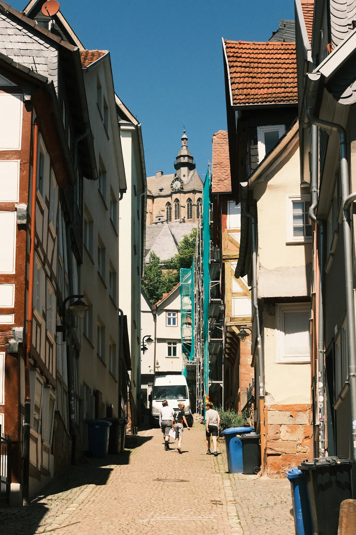 A narrow cobblestone alley in Marburg's old town, two people walking toward a church tower in the distance.