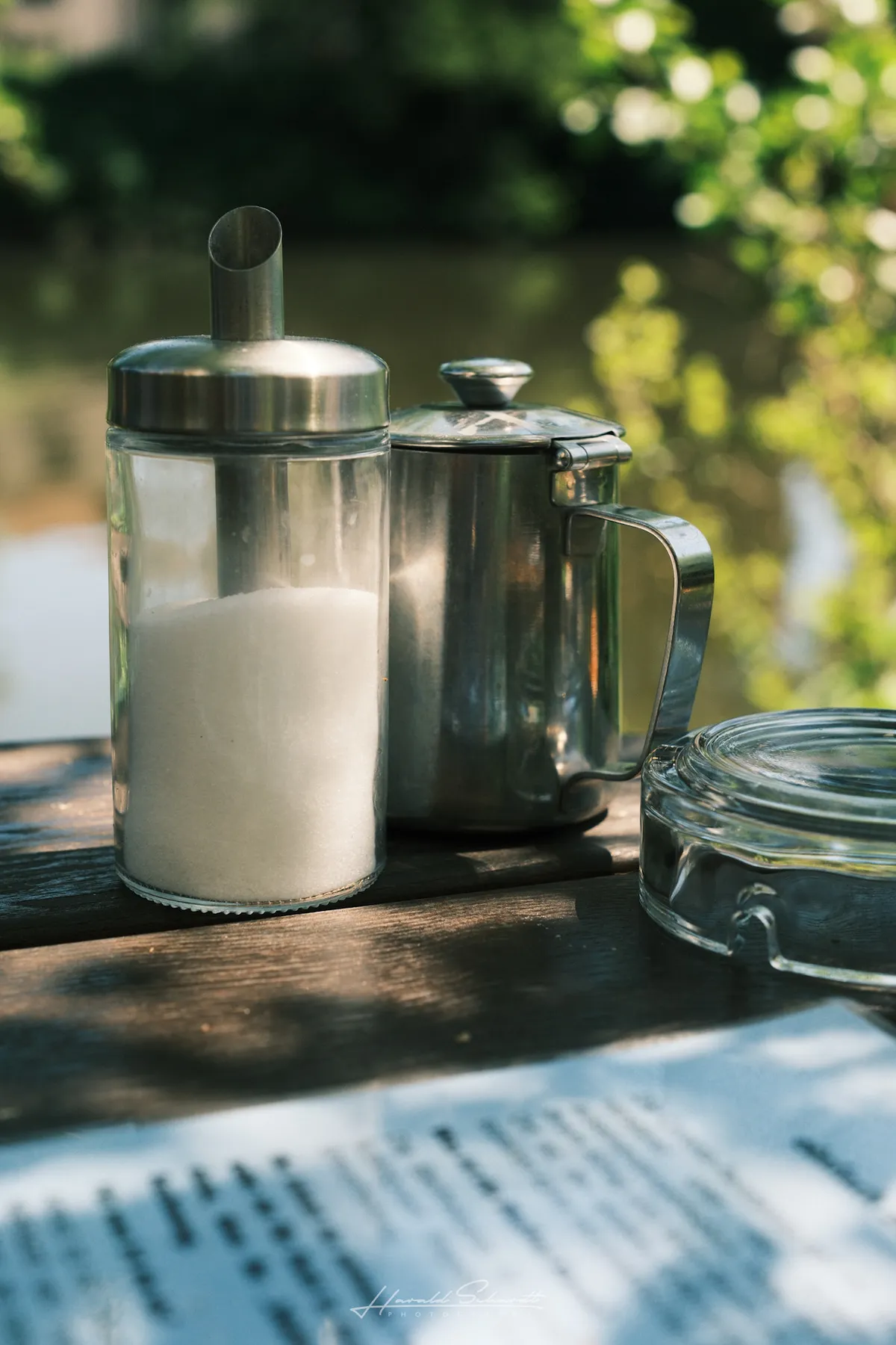 A glass sugar dispenser and stainless steel milk jug on a wooden café table, green trees blurred in the background.