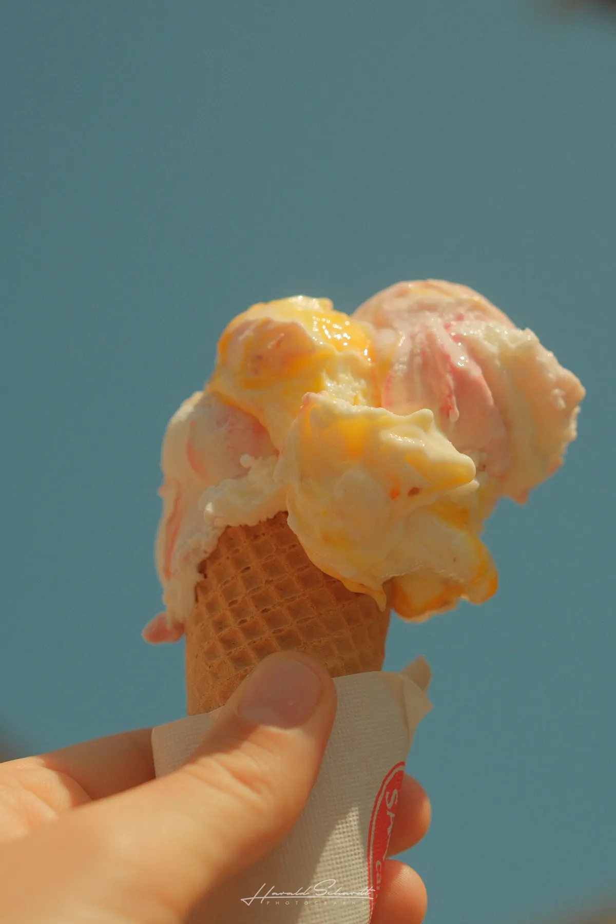 A melting ice cream cone held up against a clear blue sky, two scoops in pink and orange.