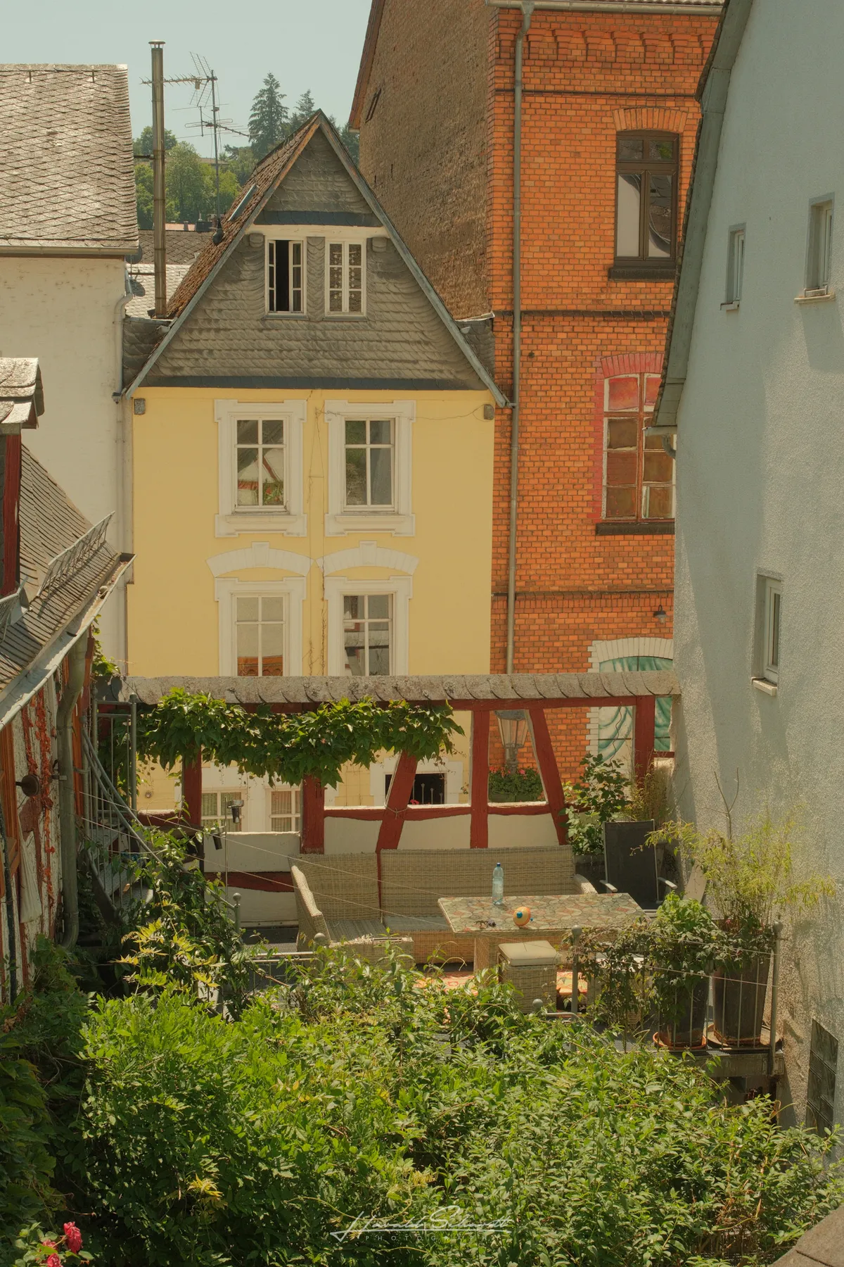 A yellow house and red brick building seen from above, with a garden terrace and pergola below.