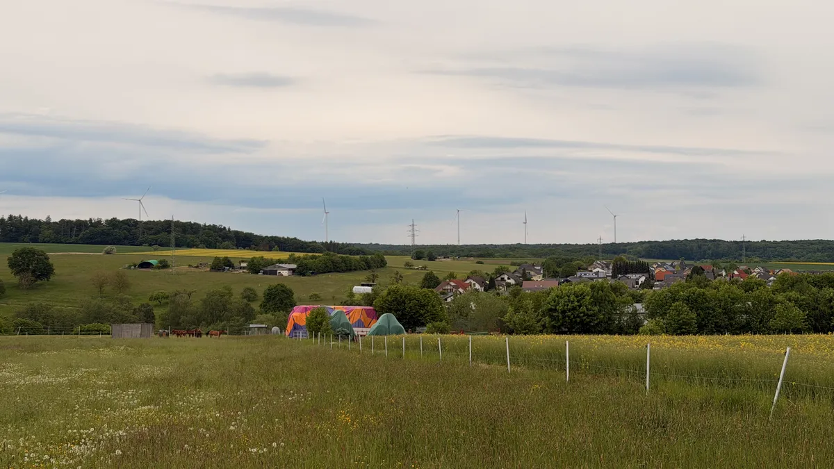 Green fields stretching to a small village. Colourful tents or inflatables visible in the middle distance, wind turbines on the horizon.