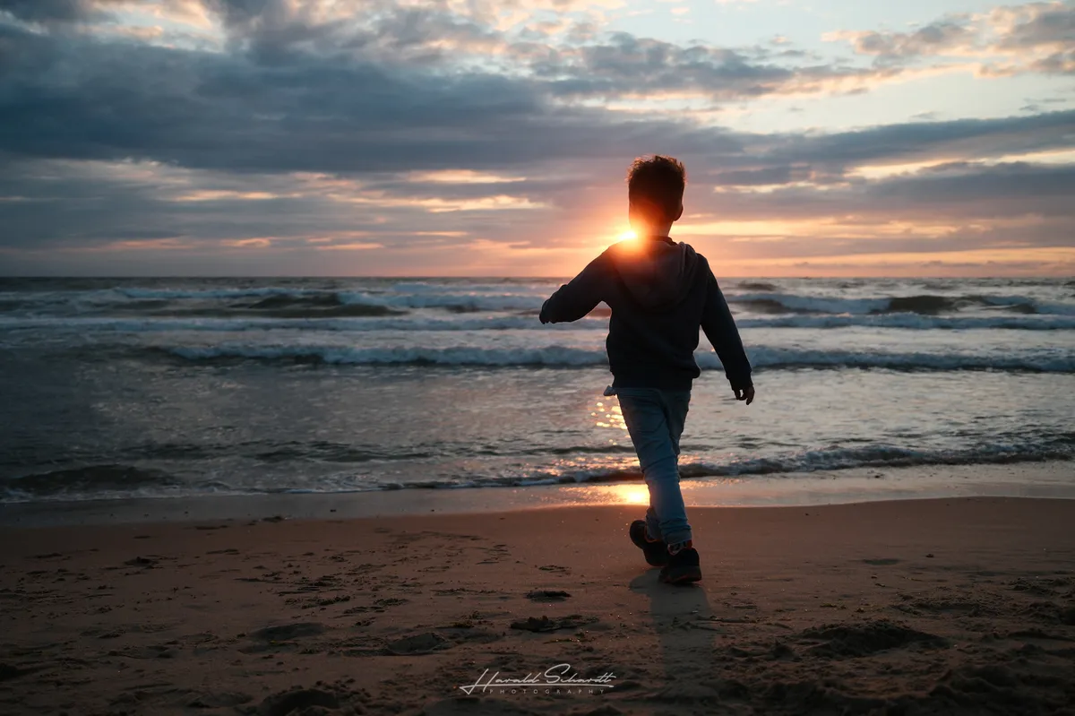A child walks toward the sea at sunset on a North Holland beach. The setting sun aligns with the child’s silhouette, creating a glowing halo effect. Dramatic cloudy sky, waves breaking in the background.