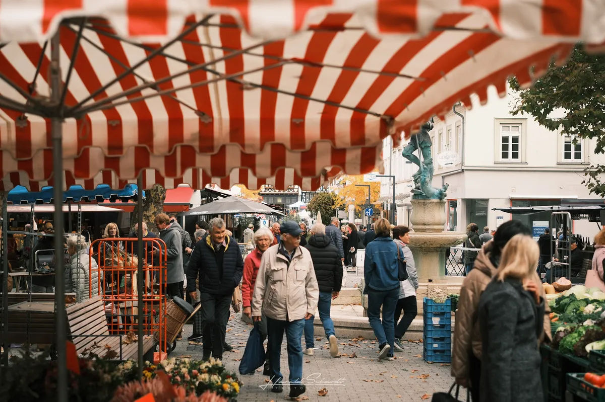 Busy outdoor market on an overcast autumn day. A red and white striped awning frames the top of the image. People in jackets and coats stroll between the stalls. A stone fountain with a figure stands in the background, flanked by white-plastered buildings. Crates of vegetables line the right edge. Fallen leaves on the cobblestones.