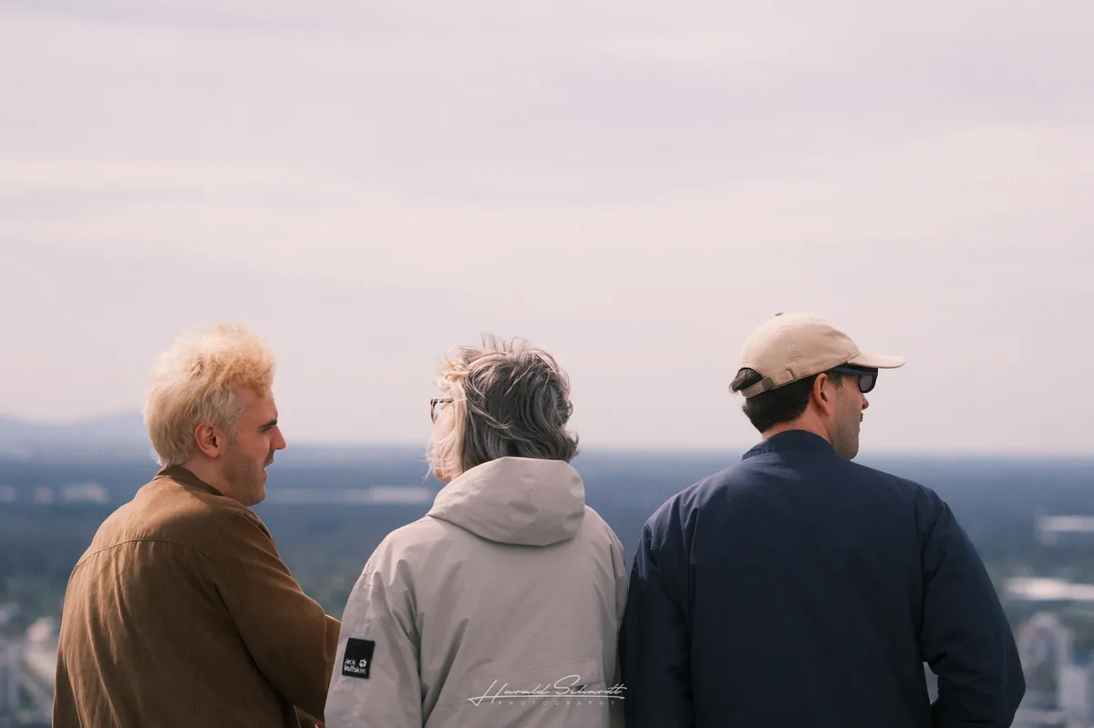 Three people seen from behind, standing close together, looking out over a wide landscape. Overcast sky, distant hills, a city spread below. The person on the left is in profile, slightly smiling.