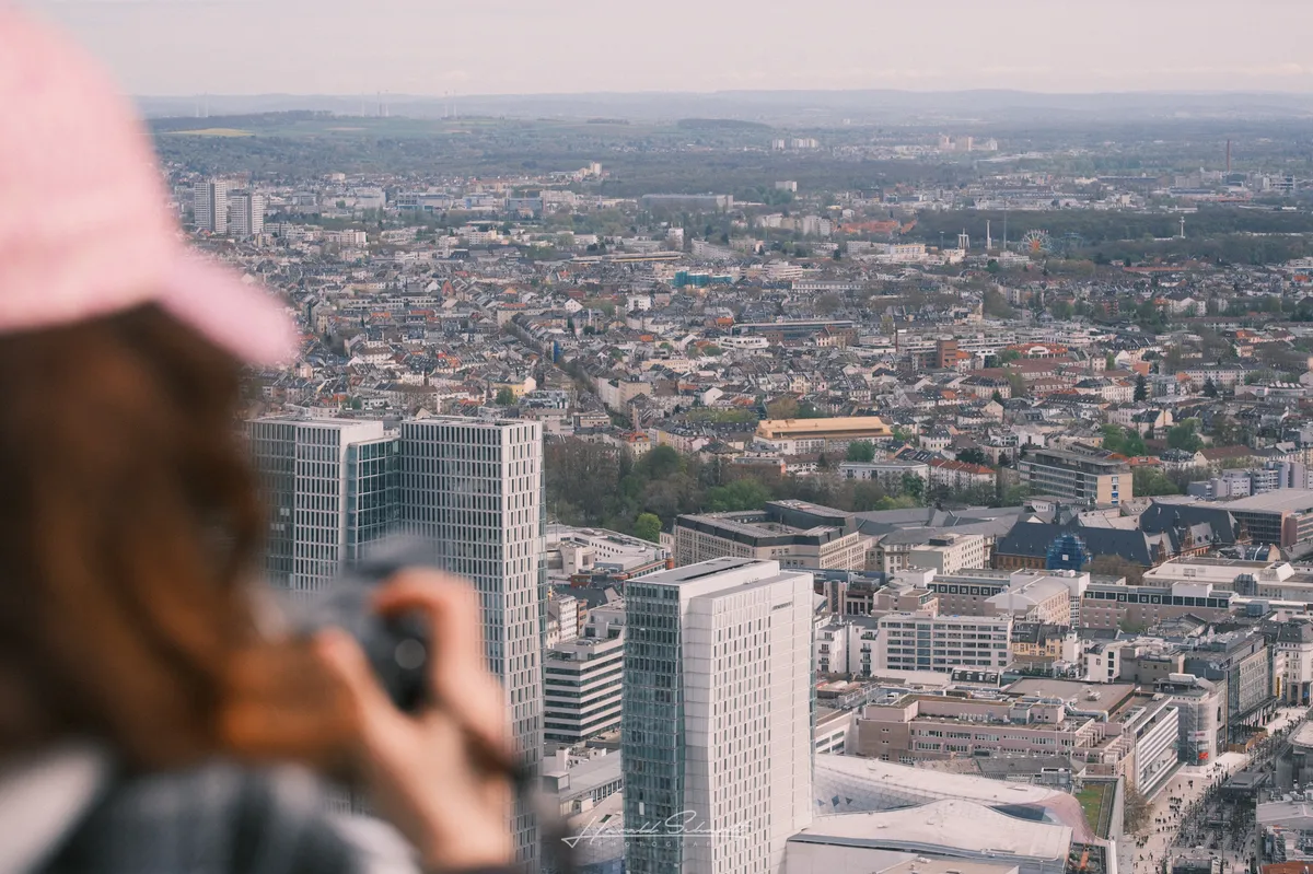 A person in a pink cap holds a camera up to photograph the city below. Out of focus in the foreground, sharp cityscape behind: rooftops, high-rises, streets stretching to the horizon.