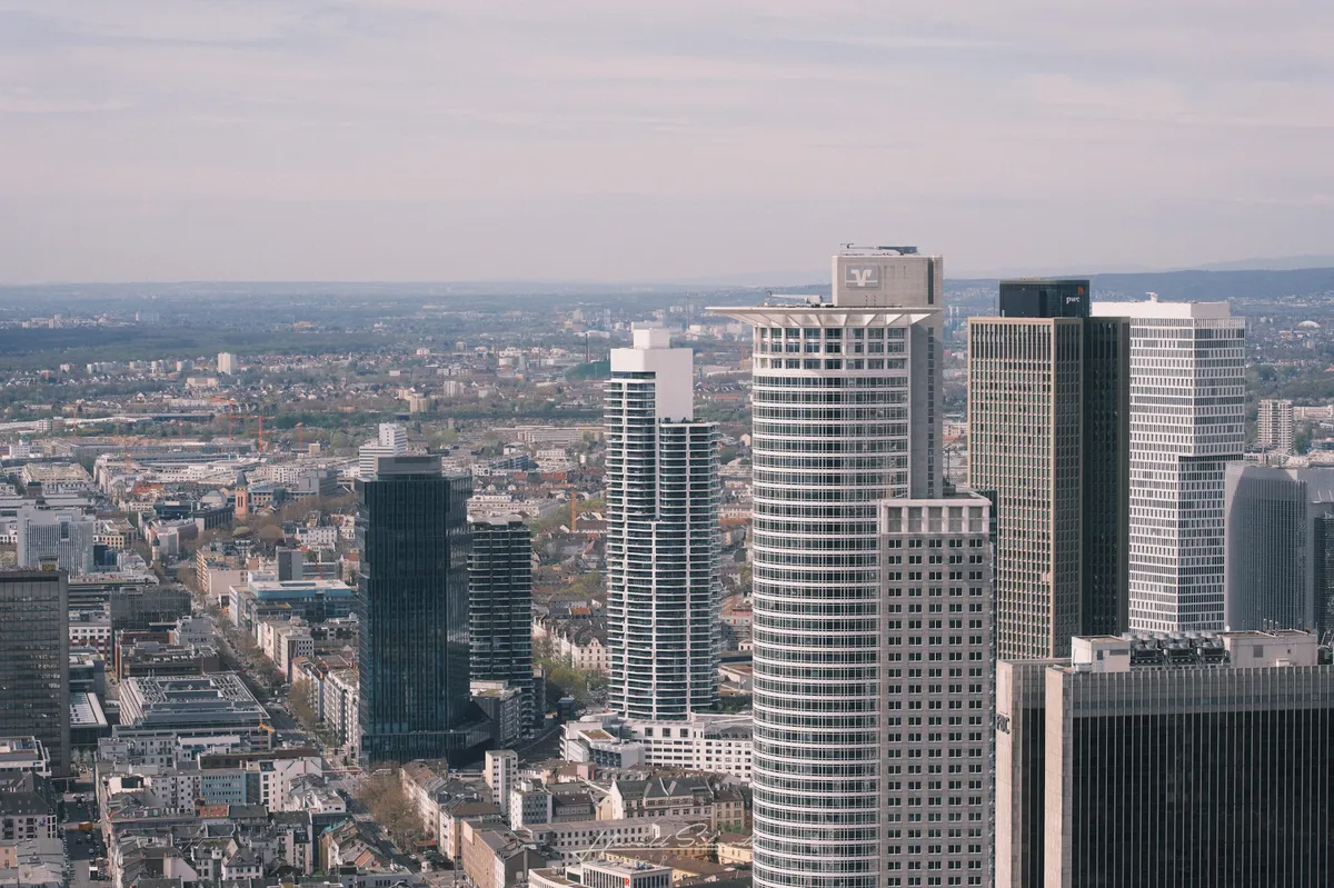 Aerial view of Frankfurt’s financial district. Several skyscrapers dominate the right side of the frame, including buildings marked Volksbank and PwC. Low-rise neighbourhoods and streets fill the background, hazy horizon beyond.