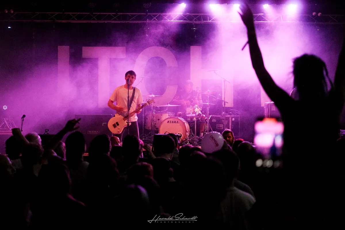Itchy live: purple haze fills the venue. Vocalist-guitarist in a white t-shirt at the mic, drummer behind. “ITCH” letters visible through the smoke on the backdrop. A silhouette with raised arm and dreadlocks in the right foreground. Tama kick drum and Meinl cymbals visible.​​​​​​​​​​​​​​​​