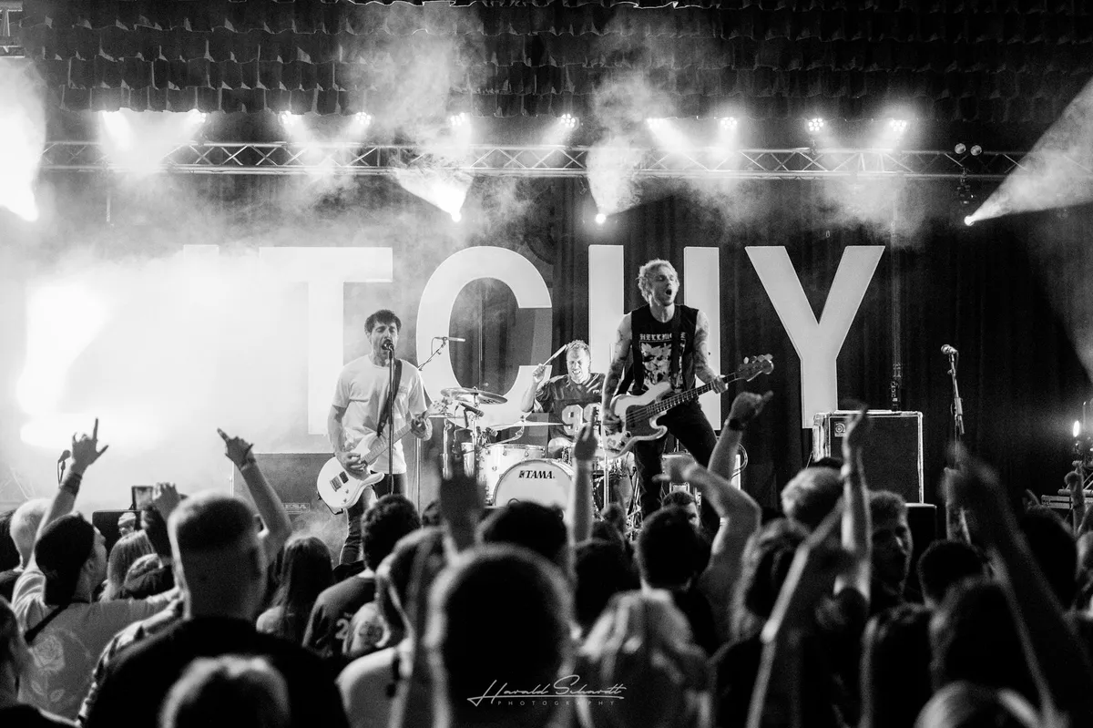 Itchy live: black and white. Three-piece band at full intensity: vocalist-guitarist left, drummer centre, bassist right with arms wide. Large “ITCHY” letters on the backdrop, partly obscured by smoke and backlighting. Crowd in front with arms raised.