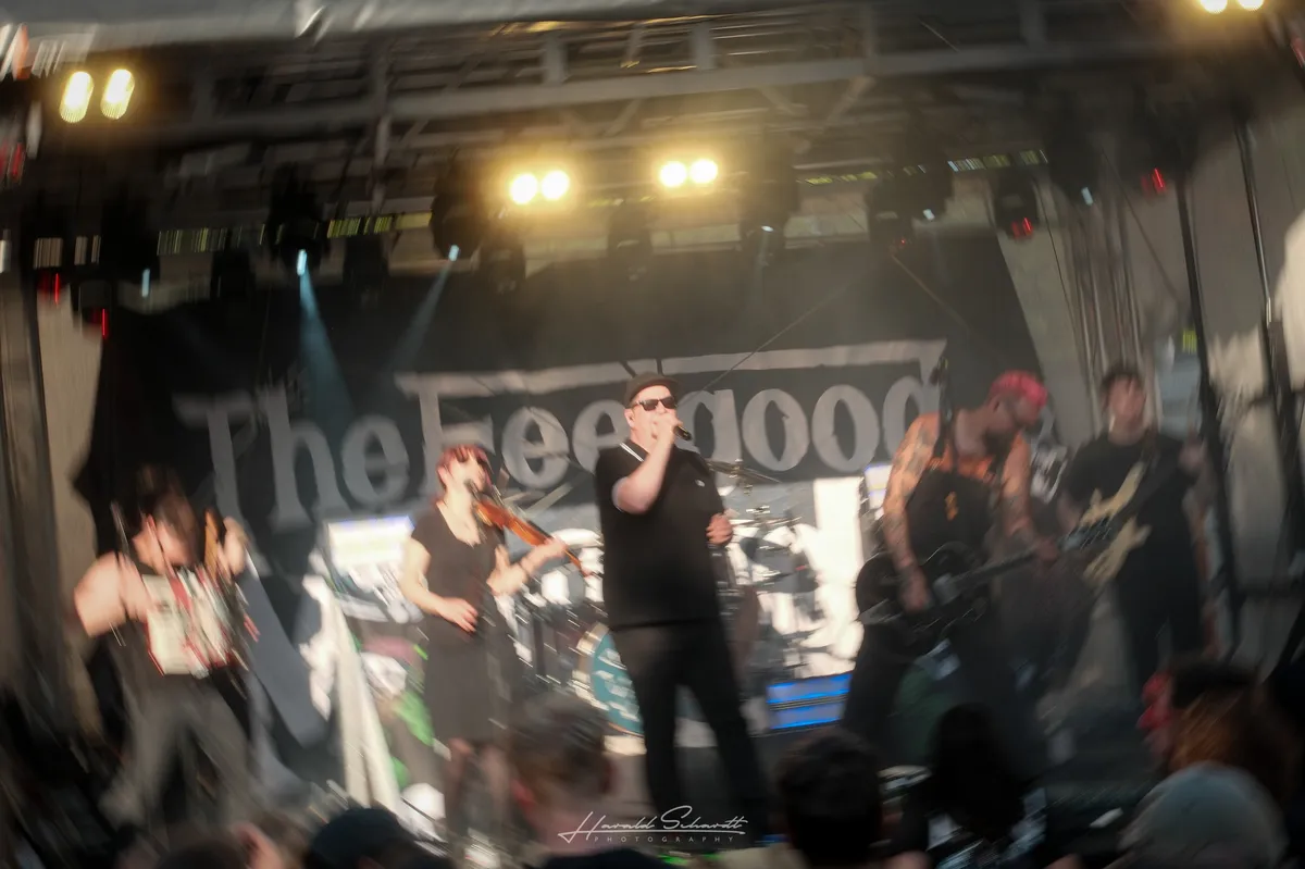 The Feelgood McLoods live: intentionally motion-blurred concert shot. A large band on stage, vocalist centre in sunglasses, violinist to his left. The band name is partially visible on the backdrop. Warm stage lights cut through the blur.