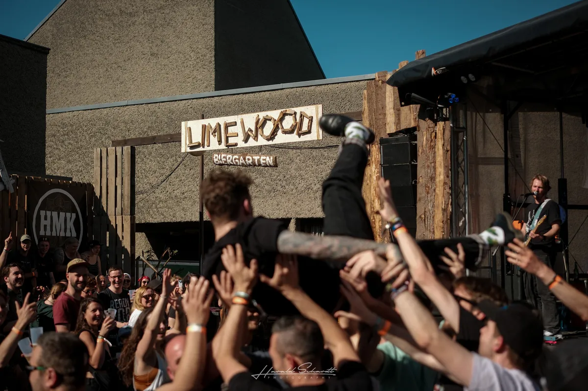 lüt live: outdoor daytime show. A crowd surfer is passed overhead by dozens of hands. The “Limewood Biergarten” sign is clearly visible on the wall behind the stage. A guitarist plays on the right edge of the frame. Bright blue sky above.