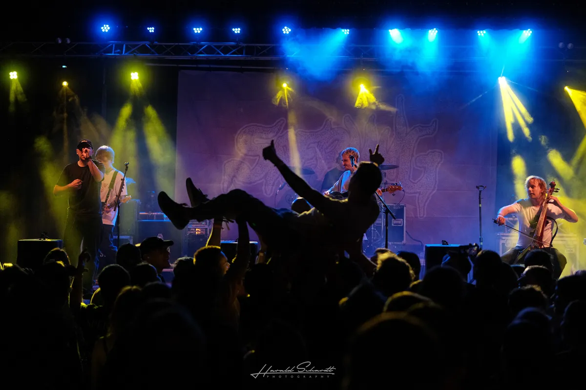 Sperling live: concert under blue and yellow stage lights with thick smoke. A crowd surfer is held up by the audience, arms raised, boots in the air. The band plays behind them: vocalist on the left, guitarist and cellist visible on stage. A large illustrated backdrop fills the background.