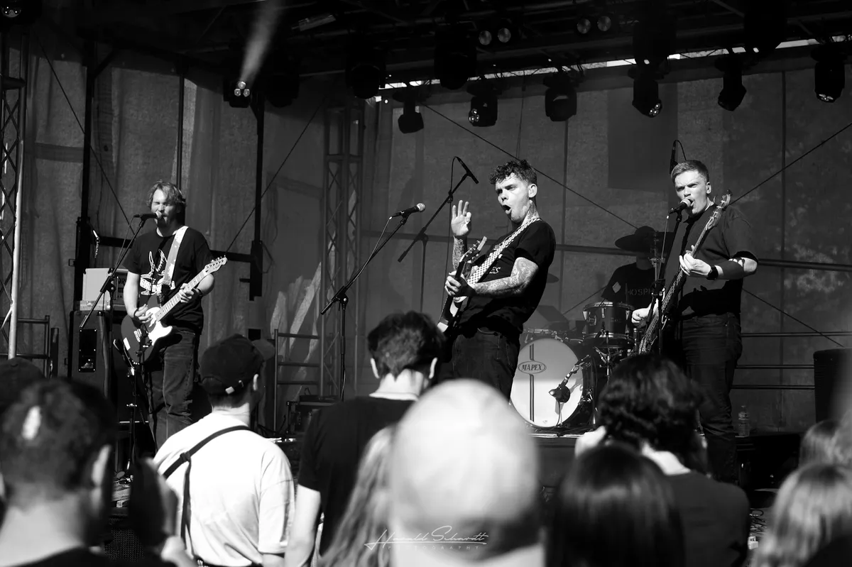 lüt live: black and white concert photo. Four-piece band on a small indoor stage. The tattooed frontman sings and raises one hand, guitar in the other. Guitarist on the left, bassist on the right, drummer partially visible behind. Audience heads in the foreground.