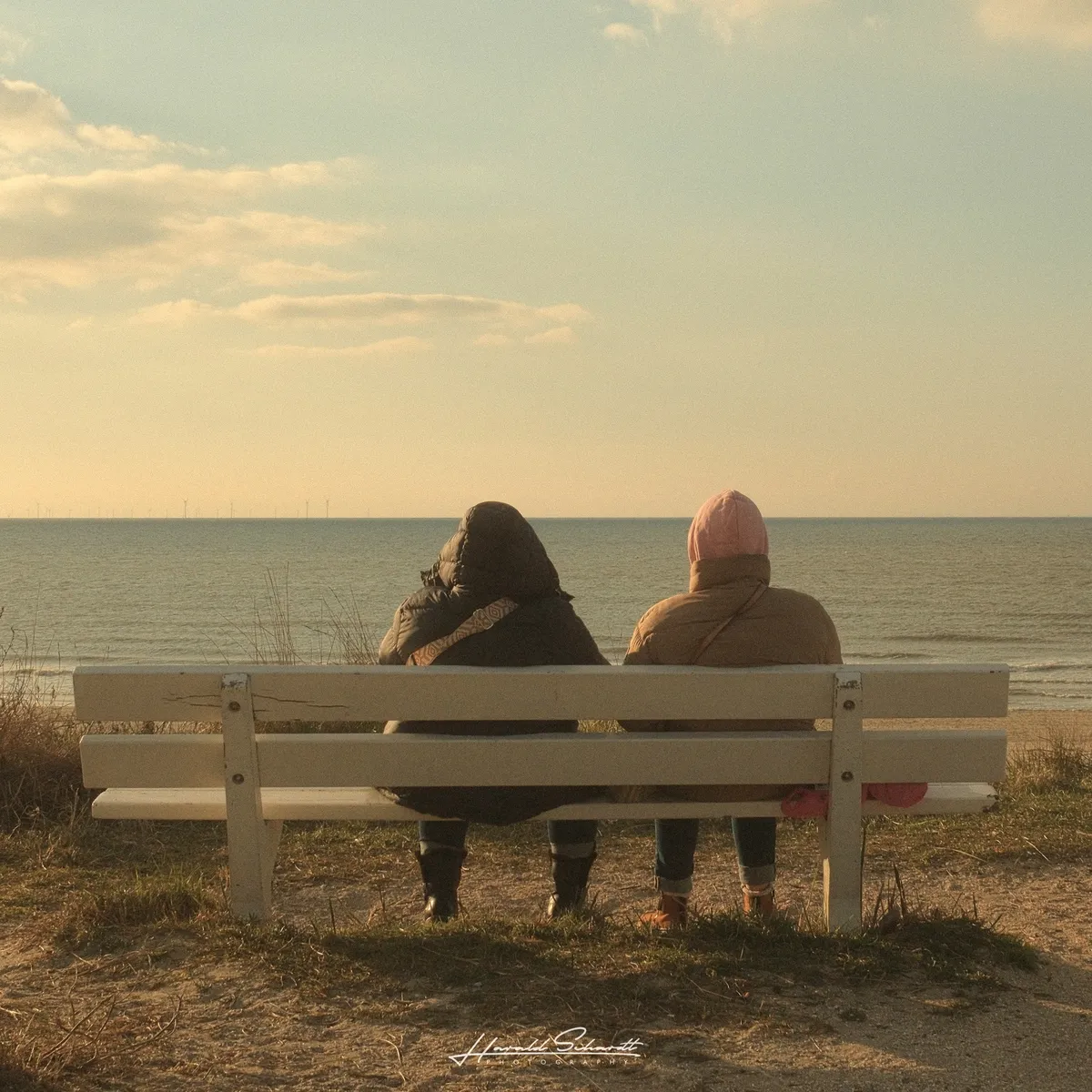 Two people sitting side by side on a white bench, seen from behind, looking out over a calm body of water. Warm evening light, clouds on the horizon.