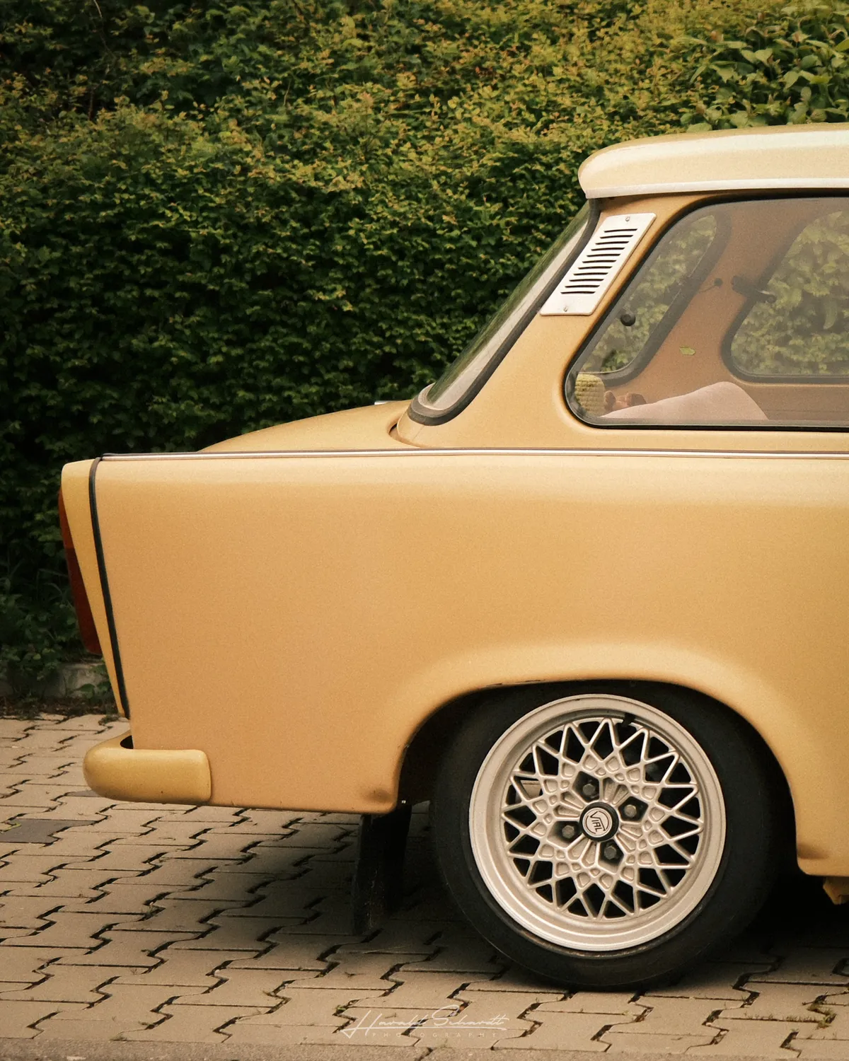 Rear quarter of a lowered golden Trabant on cobblestone. BBS-style mesh wheels, green hedge in background.