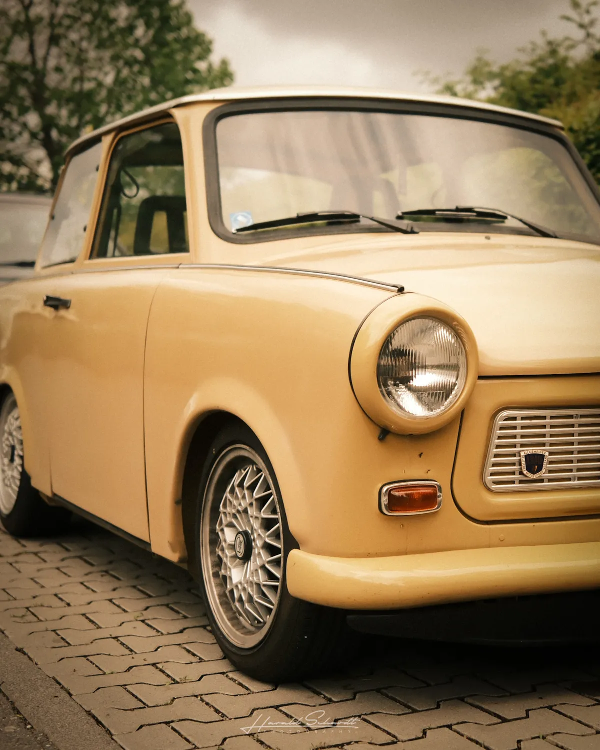 Front three-quarter view of a lowered Trabant. Round headlights, mesh wheels, pale yellow bodywork. Overcast sky above.