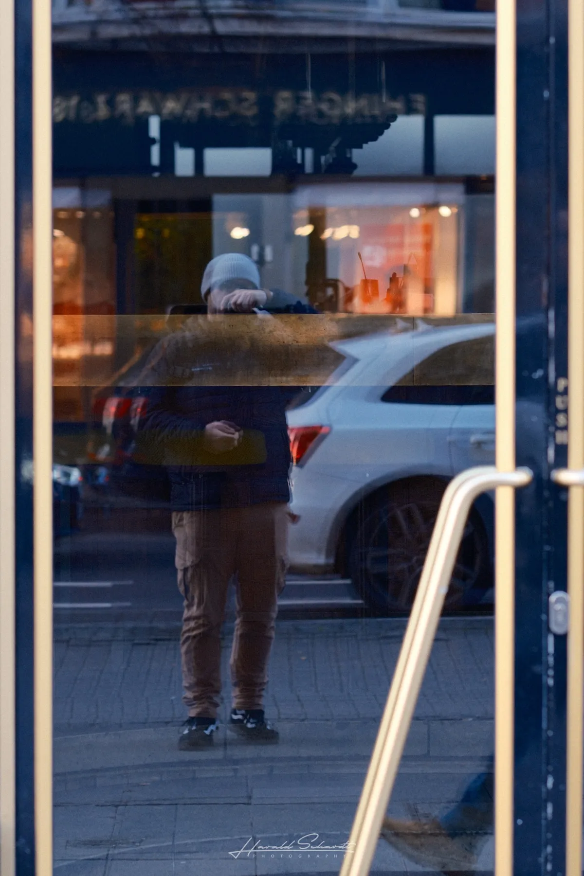 A person in a beanie and dark jacket photographs themselves in the reflection of a shop’s glass door. Street and parked cars visible in the background.​​​​​​​​​​​​​​​​