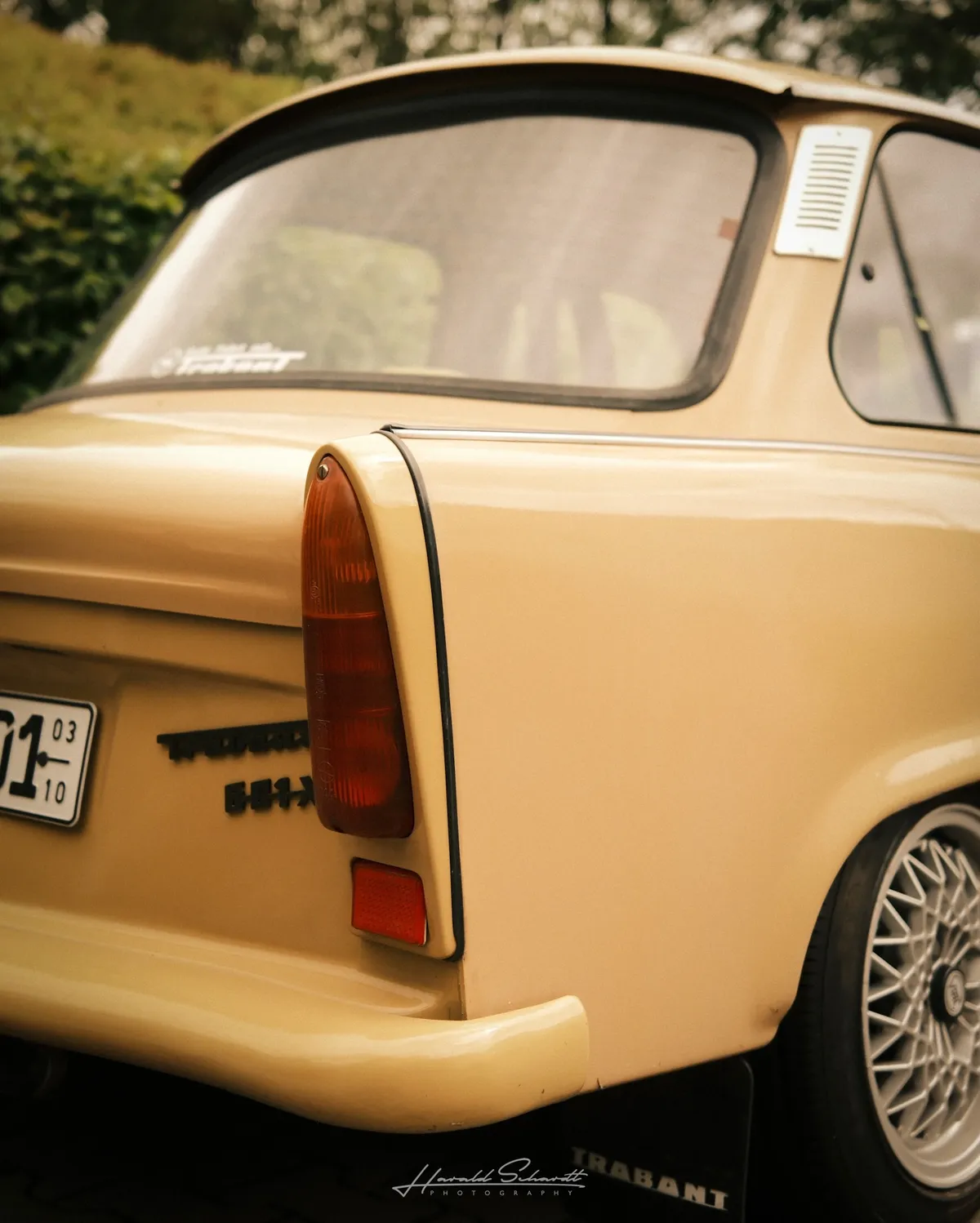 Rear three-quarter view of a lowered Trabant 601. Tail lights, badge, and partial licence plate visible. Mesh wheels, hedge in background.
