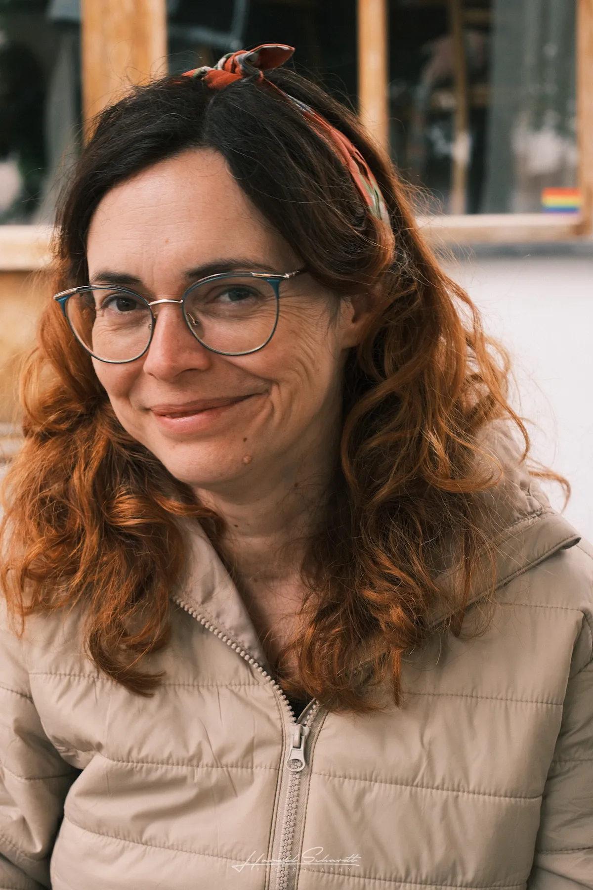 A woman with curly reddish hair and glasses, smiling slightly. Outdoor café background, soft light.