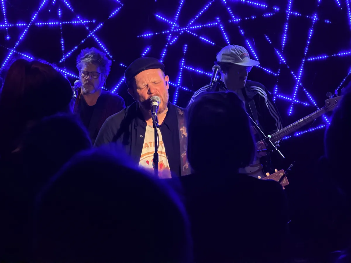 Matze Rossi live: singing with eyes closed at a standing microphone, black beret, tattoos visible on his neck. Two band members behind him, blue LED star-pattern backdrop. Audience silhouettes in the foreground.