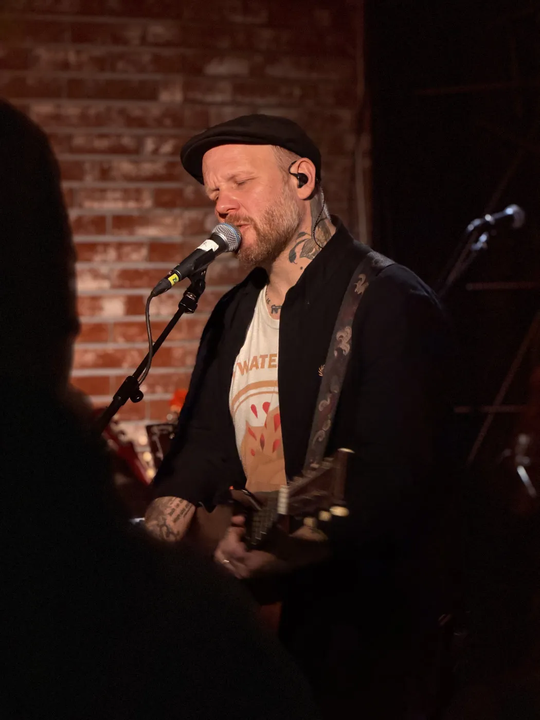Matze Rossi live: close-up portrait at the mic, eyes closed, flat cap, neck tattoos, in-ear monitor visible. Brick wall background, warm stage lighting. Acoustic guitar strap across his chest.