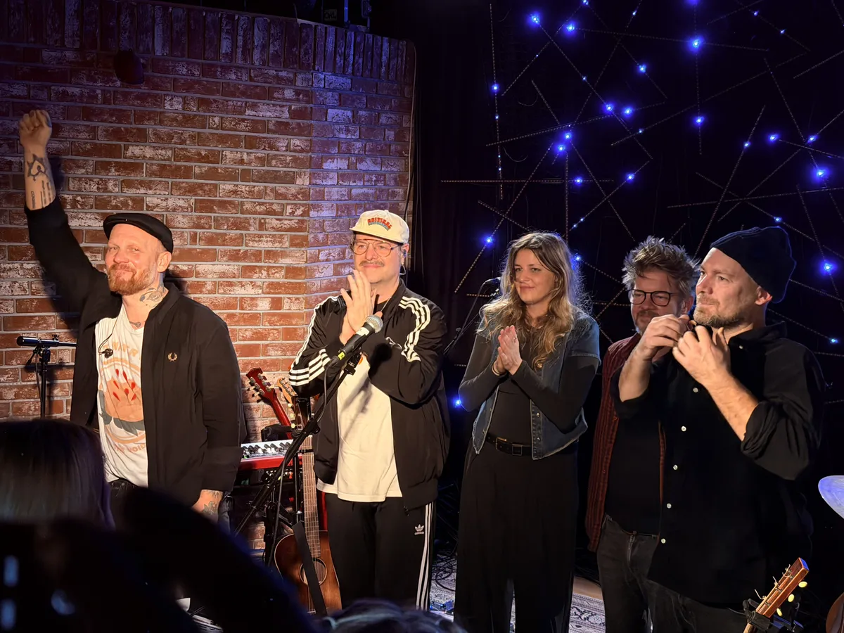 Matze Rossi and his band taking a bow after the show. Five musicians on stage in front of a brick wall with blue LED star decorations. Matze on the far left, fist raised, tattooed arm in the air. The rest of the band applauding beside him.
