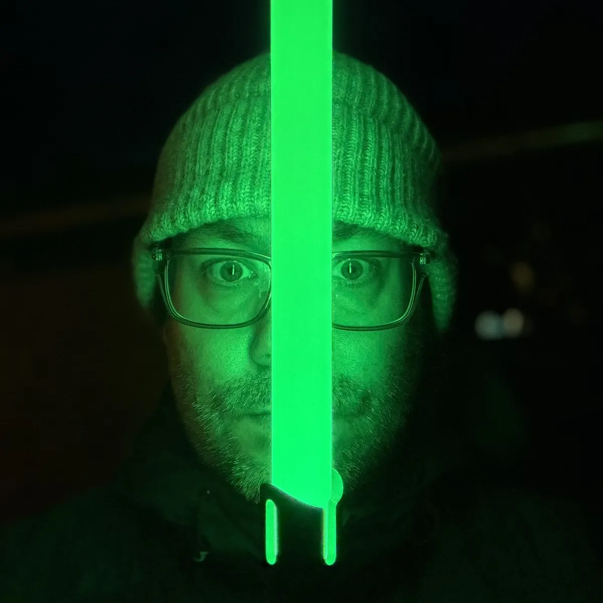 A person wearing a knit beanie and glasses, face lit by a green lightsaber held vertically in front of them. Dark background, strong green glow.