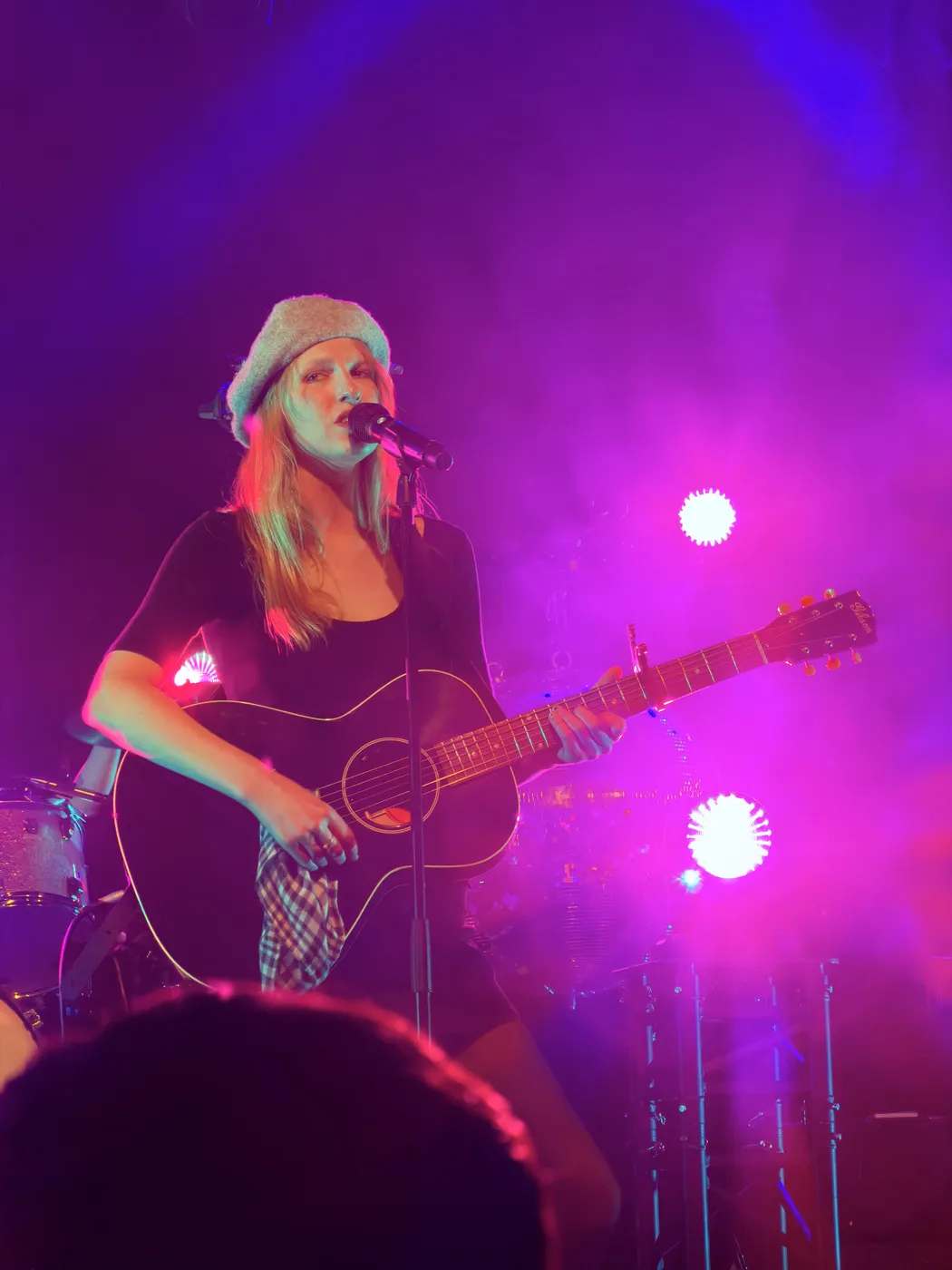 Audrey Hobert live: standing at the mic with an acoustic guitar, grey bucket hat. Pink and magenta stage lighting, drummer partially visible behind her.