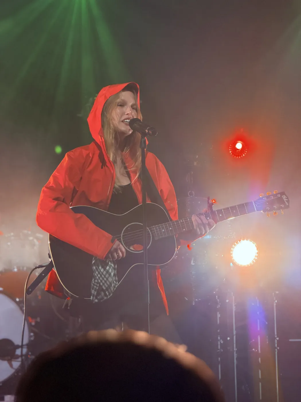 Audrey Hobert live: playing acoustic guitar in a red hooded jacket, singing at the mic. Warm orange stage light to the right, green light in the background. Crowd heads visible in the foreground.