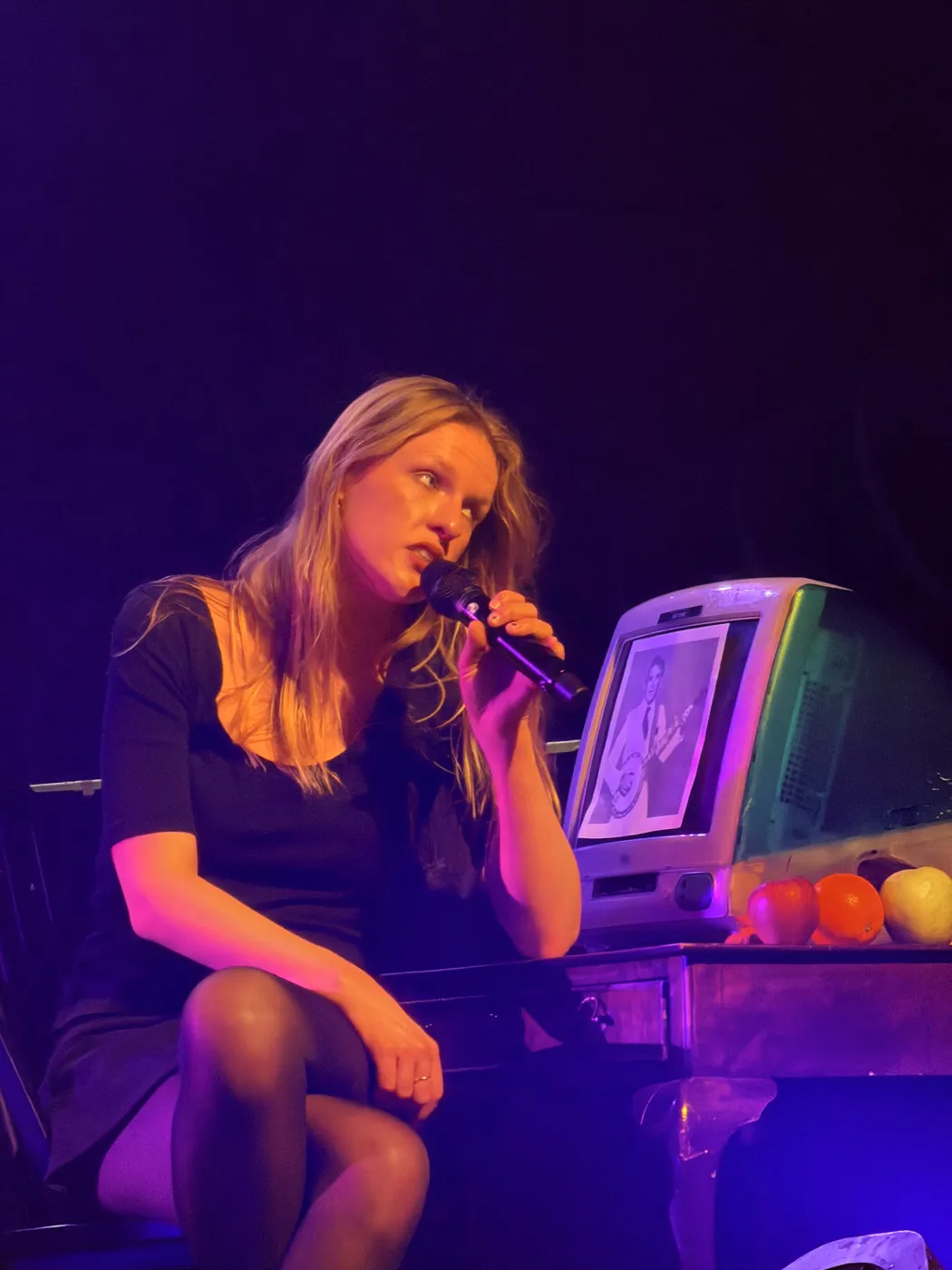 Audrey Hobert live: seated on stage, singing into a handheld microphone. Black dress, long blonde hair. A retro CRT monitor and coloured balls visible in the background. Dark, intimate venue.