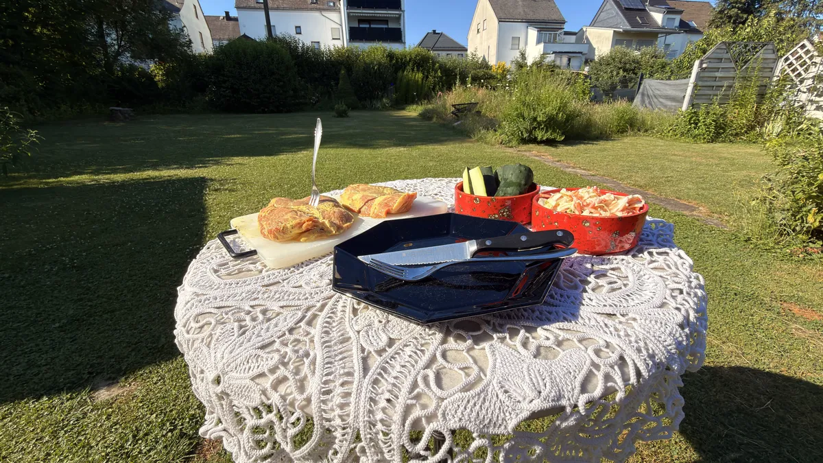 A small garden table prepared for a solo barbecue, with marinated steaks, salad, vegetables, and evening sunlight stretching across the lawn.