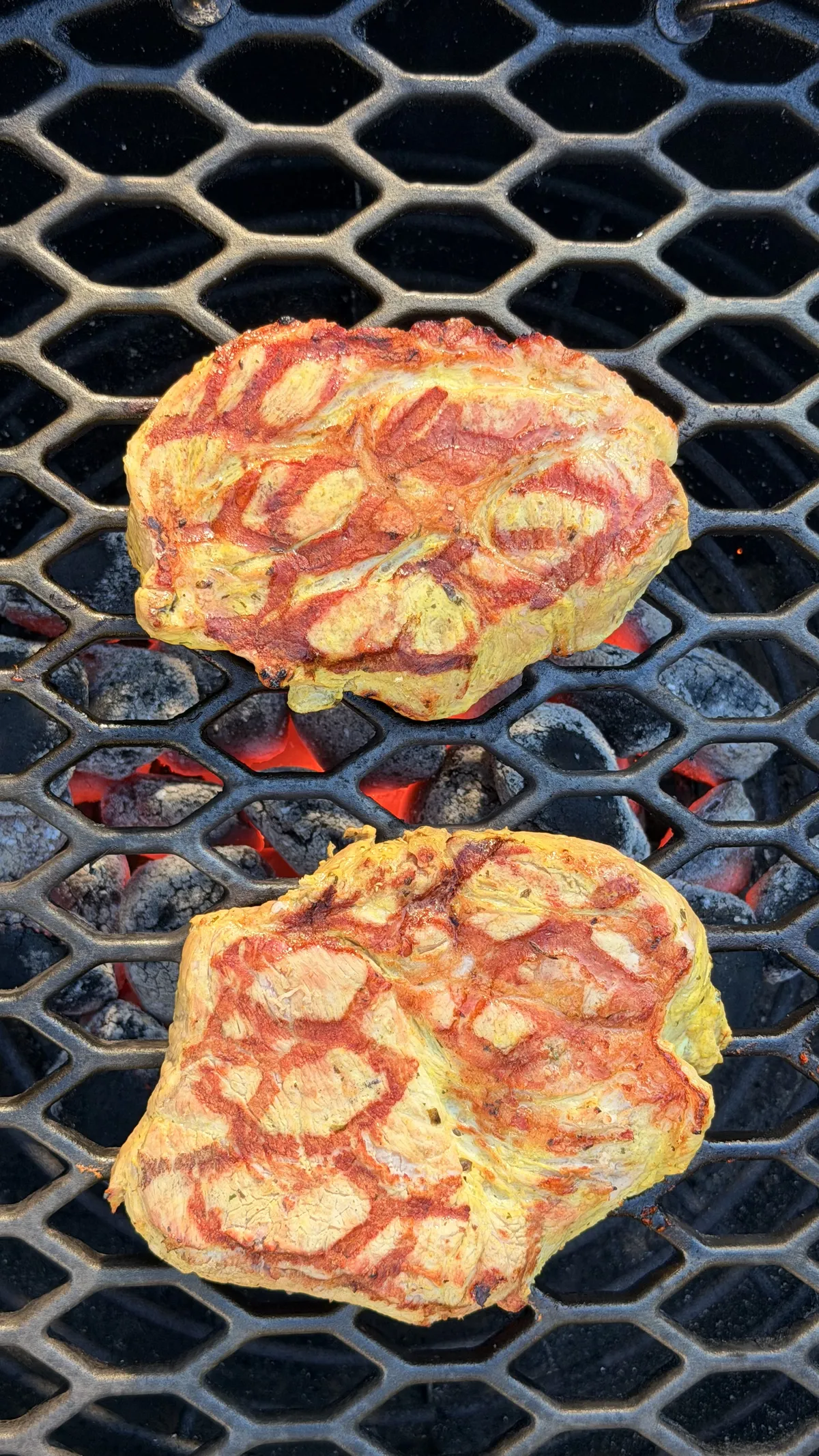 Two marinated pork neck steaks grilling over glowing charcoal on a barbecue, viewed from above.