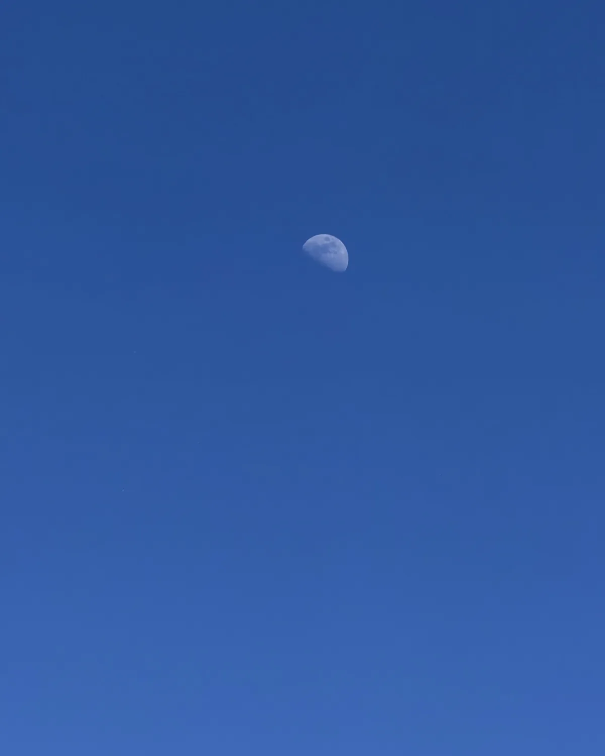 Half moon in a clear blue daytime sky, photographed handheld with a mobile phone.