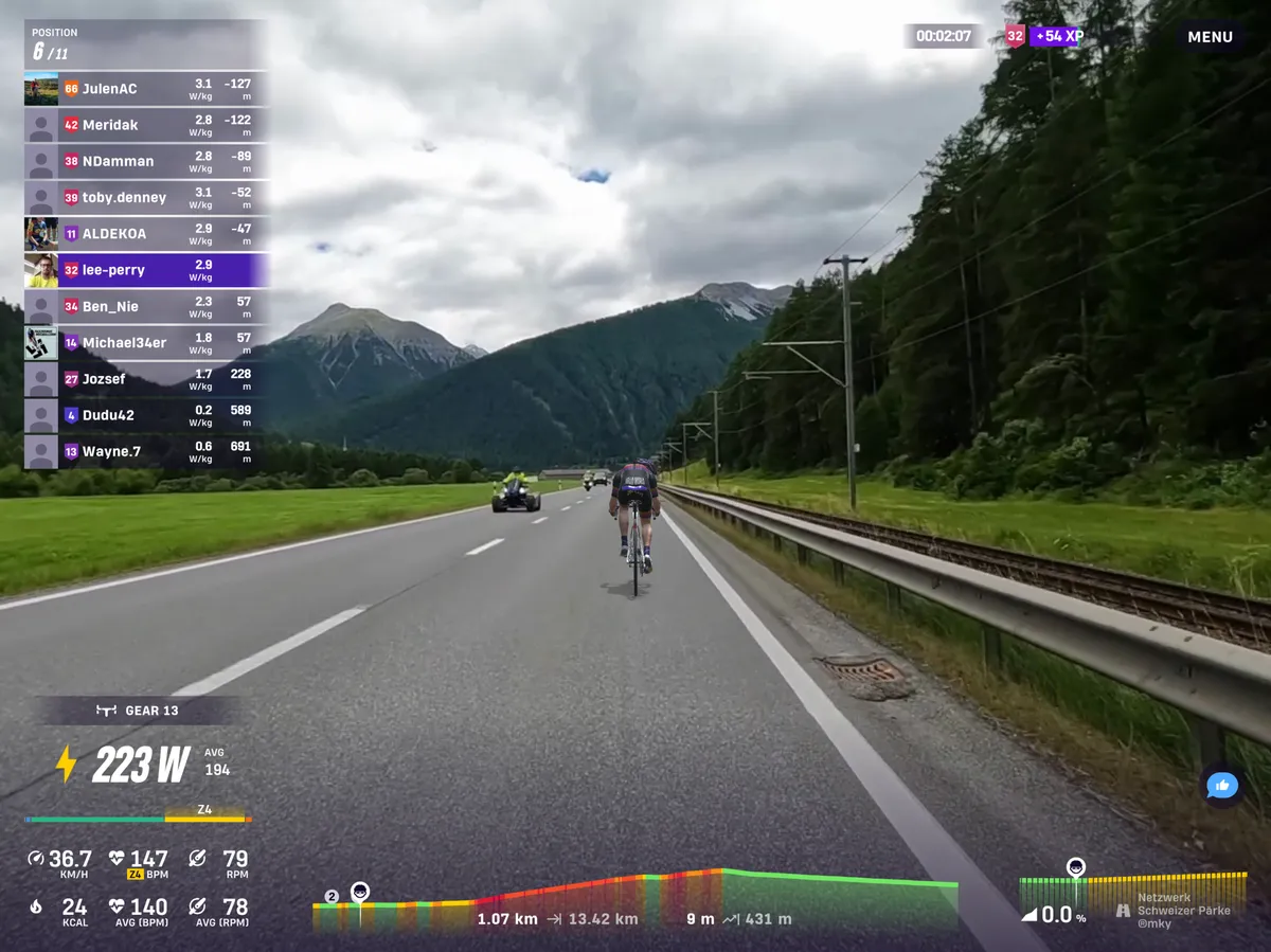 A cyclist's avatar riding through a green valley on Rouvy with mountains and grey skies beyond