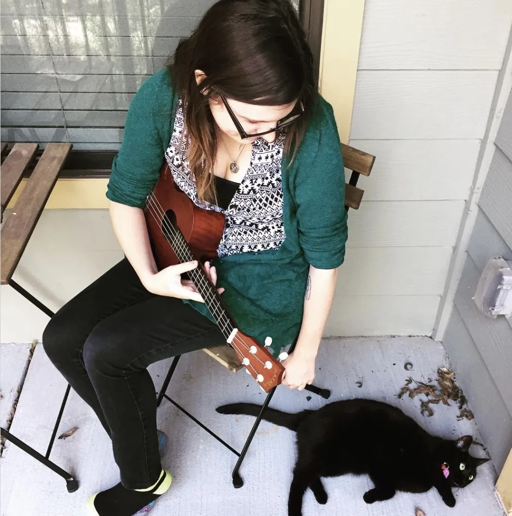 Megan sitting on an outdoor chair holding a ukulele, a black cat laying on the ground next to her