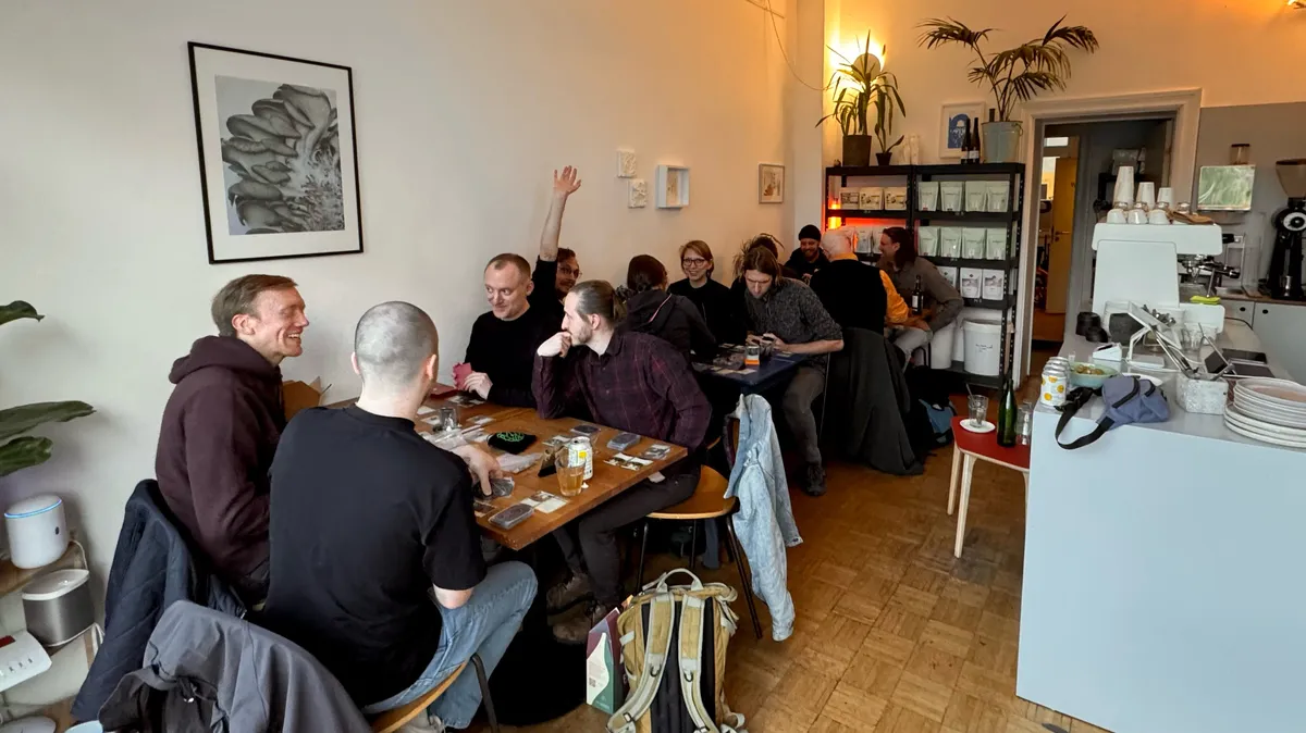 Group photo at the tables. There are 12 players sitting across 3 tables in a coffee shop. Each table has players intently doing initial setup, planning, and strategising.