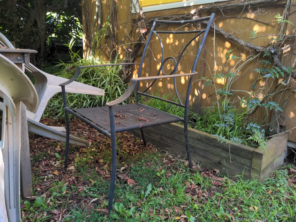 a rusty metal chair, covered in dirt and leaves, sits at a rusty table, surrounded by unkempt grass, in front of a rustic cabin