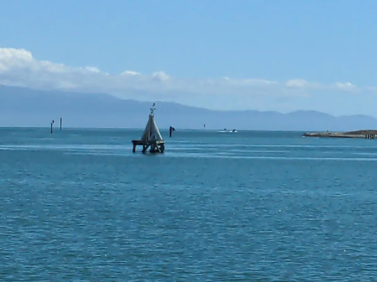 a sunny day overlooking the ocean. no waves and a single small structure sits in the middle where a tepee looking buoy thing rests on top.